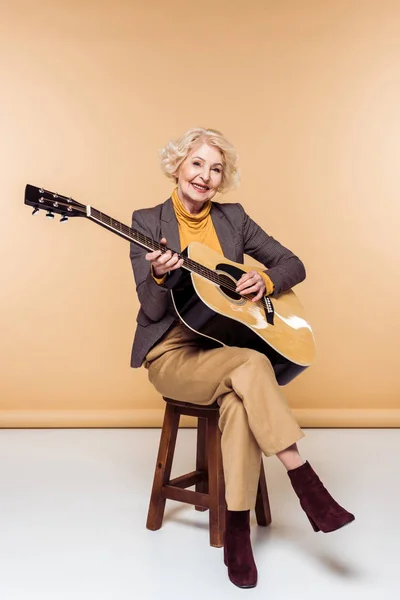 Sonriente mujer mayor con estilo sentado en la silla y tocando en la guitarra acústica - foto de stock