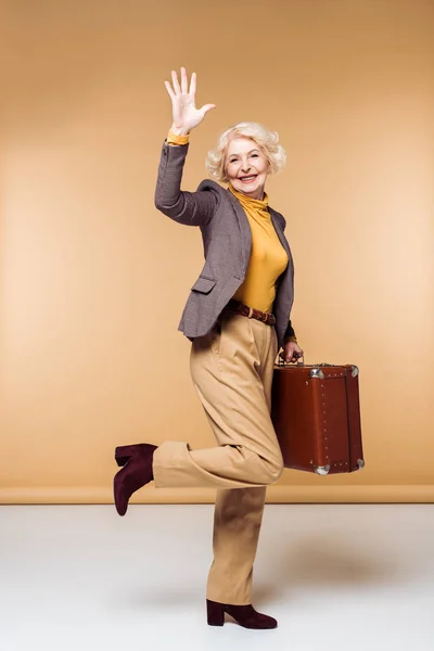 Stylish senior female traveler waving by hand and holding vintage suitcase — Stock Photo