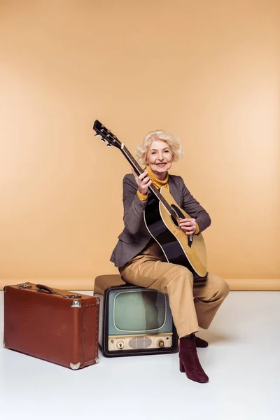 Senior woman with acoustic guitar sitting on vintage tv near old suitcase — Stock Photo