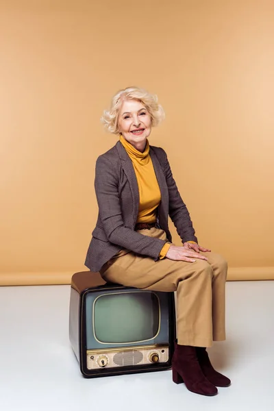 Улыбающаяся стильная пожилая женщина сидит на винтажном телевизоре — стоковое фото