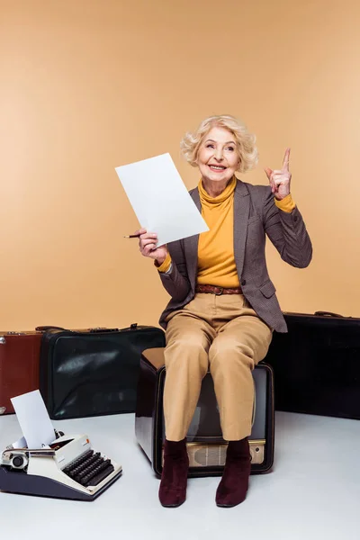 Улыбающаяся стильная пожилая женщина с поднятым пальцем, держащая бумагу и сидя на винтажном телевизоре рядом с пишущей машинкой и чемоданами — стоковое фото