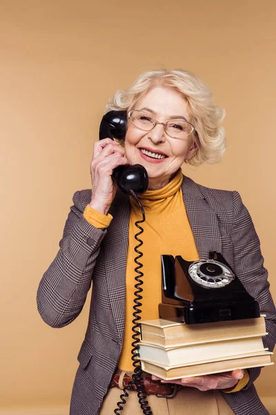 Happy fashionable senior woman in eyeglasses talking on rotary phone and holding stack of books — Stock Photo