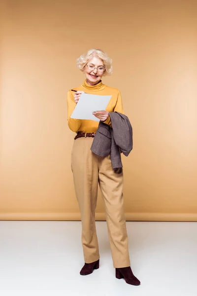 Mulher sênior elegante em óculos segurando jaqueta e escrevendo em papel — Fotografia de Stock