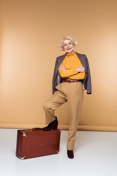 Модная пожилая женщина-туристка в пиджаке на плече держит ногу на винтажном чемодане — стоковое фото