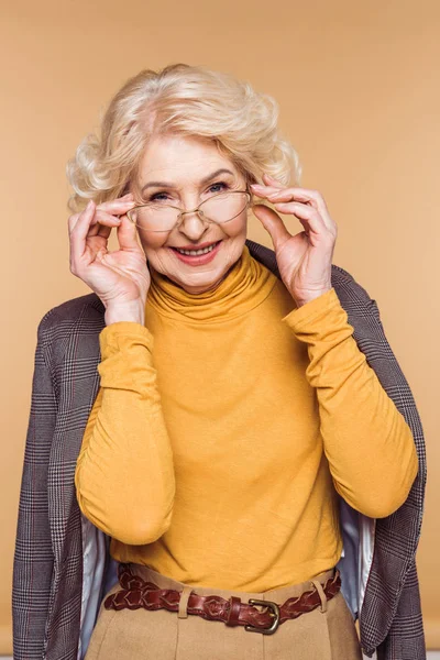 Улыбающаяся модная пожилая женщина в пиджаке на плечах регулирует очки изолированные на бежевом фоне — стоковое фото