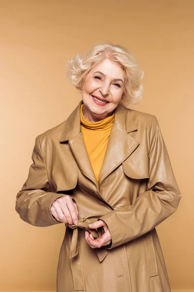 Sonriente mujer senior de moda atando cinturón de abrigo de trinchera de cuero aislado sobre fondo beige - foto de stock