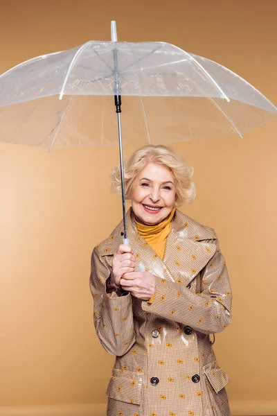 Stylisch lächelnde Seniorin im Trenchcoat mit Regenschirm auf beigem Hintergrund — Stockfoto