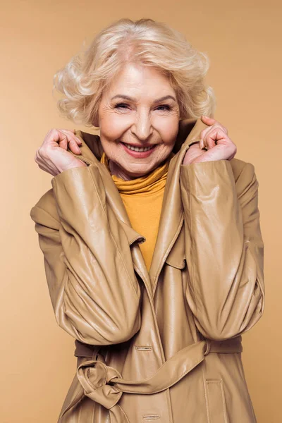 Усміхнена стильна старша жінка позує і тримає комір траншеї пальто — стокове фото