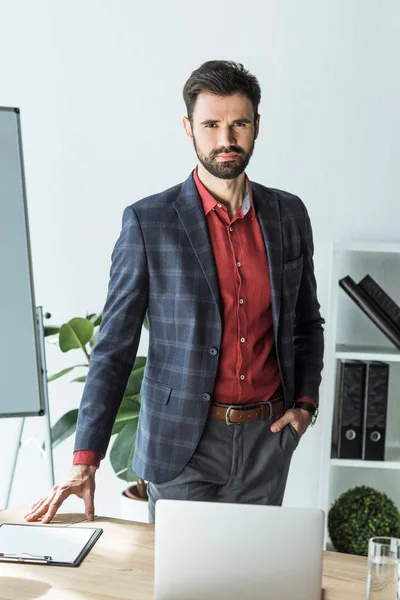 Attractive young businessman standing next to his workplace at office — Stock Photo