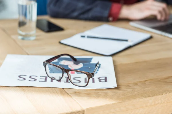 Nahaufnahme einer Brille, die auf einer Zeitung liegt, mit einem verschwommenen Geschäftsmann, der am Hintergrund arbeitet — Stockfoto