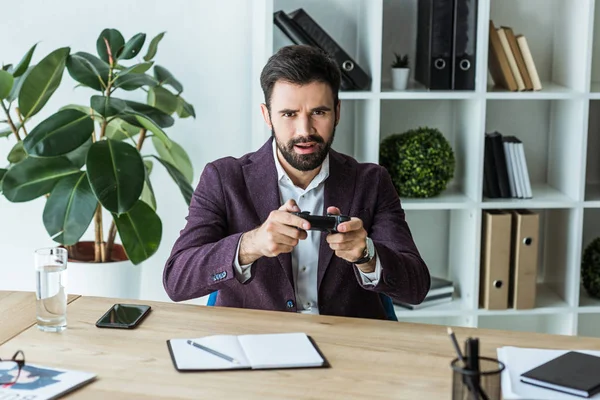 Beau jeune homme d'affaires avec manette de jeu regardant la caméra tout en étant assis sur le lieu de travail — Photo de stock