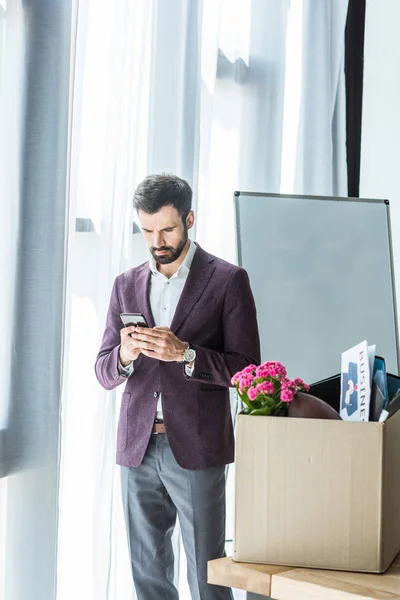 Fired young businessman using smartphone near box of personal stuff at office — Stock Photo