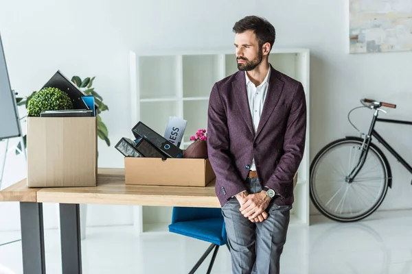 Pensativo joven hombre de negocios con cajas de cosas personales apoyado de nuevo en el escritorio en la oficina - foto de stock