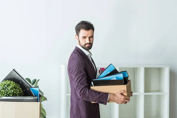 Seitenansicht eines gefeuerten jungen Geschäftsmannes, der Kiste mit persönlichen Sachen im Büro trägt — Stockfoto