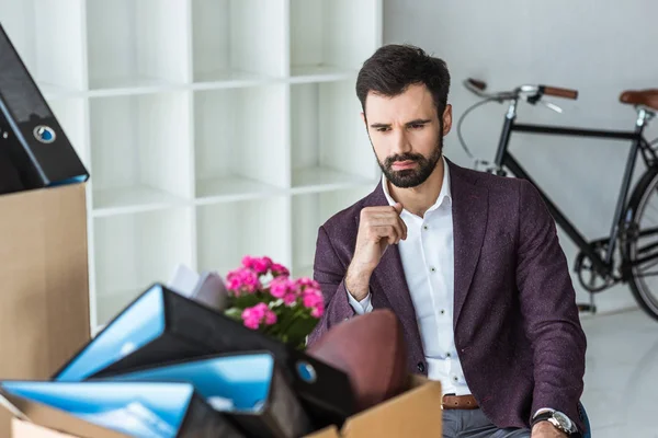 Fired young businessman sitting in office and looking at box of personal stuff — Stock Photo