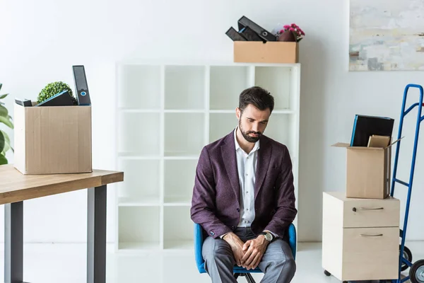 Depressed fired businessman sitting on chair in office with boxes of personal stuff — Stock Photo