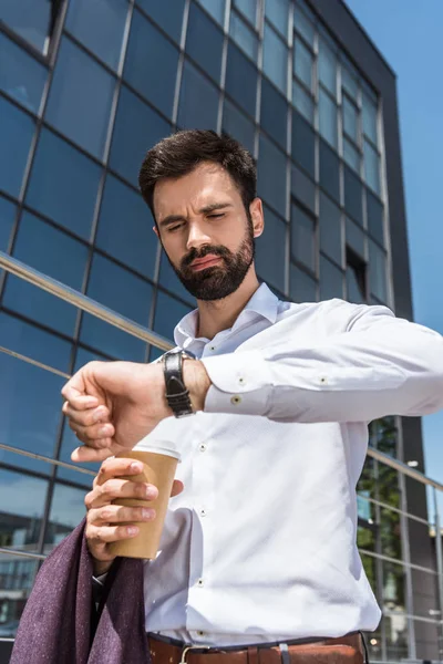 Blick von unten auf den hübschen jungen Geschäftsmann mit Coffee to go auf die Armbanduhr — Stockfoto