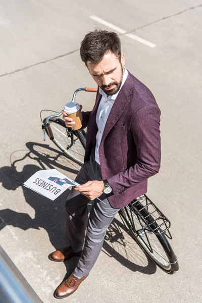 Visão de alto ângulo de belo jovem empresário com café para ir e jornais inclinados para trás na bicicleta — Fotografia de Stock