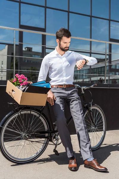Молодой менеджер с коробкой личных вещей на велосипеде смотрит на наручные часы — стоковое фото
