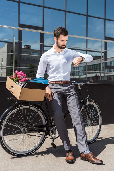 Joven gerente con caja de cosas personales en bicicleta mirando reloj de pulsera - foto de stock