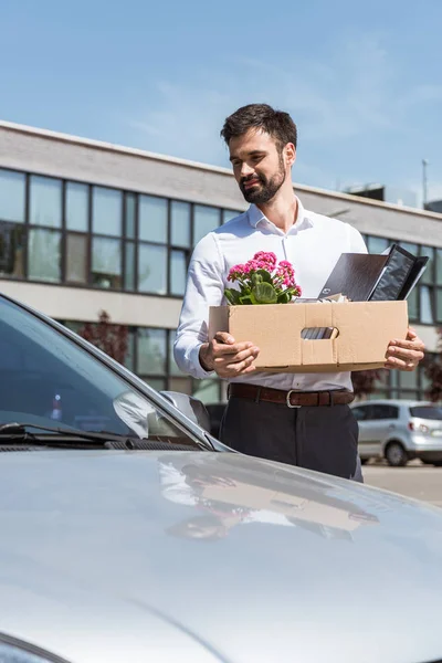 Heureux jeune gestionnaire avec boîte de choses personnelles debout près de la voiture sur le parking — Photo de stock