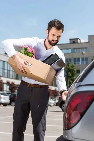 Junger Manager mit Kiste voller persönlicher Sachen öffnet Kofferraum auf Parkplatz — Stockfoto