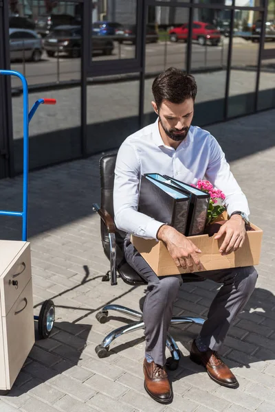 Giovane manager triste licenziato con scatola di roba personale seduta sulla sedia all'aperto — Foto stock