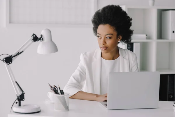Привлекательный африканский американский бизнесмен сидит за столом с ноутбуком и смотрит в сторону в офисе — стоковое фото