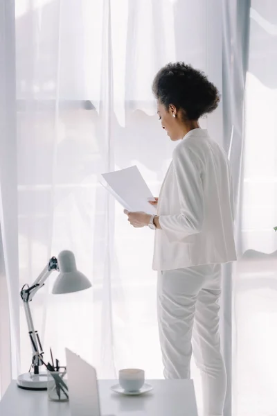 Atractiva mujer de negocios afroamericana en traje blanco leyendo documentos en la oficina - foto de stock