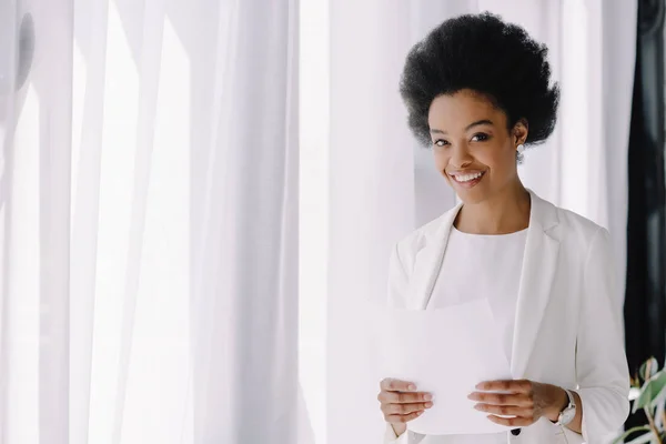 Atractiva mujer de negocios afroamericana sonriente sosteniendo documentos en el cargo - foto de stock
