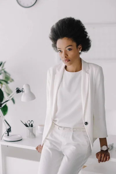 Atractiva mujer de negocios afroamericana apoyada en la mesa y mirando a la cámara en la oficina — Stock Photo