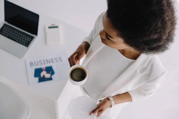 Високий кут зору приваблива афроамериканська бізнес-леді тримає чашку кави біля столу в офісі — стокове фото
