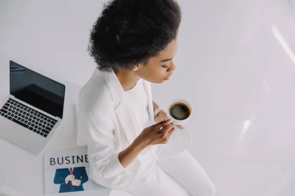 High angle view of attractive african american businesswoman sitting on table with cup of coffee in office — Stock Photo