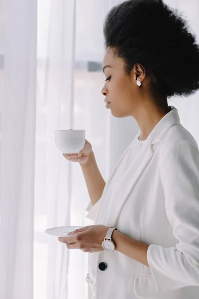 Atractiva mujer de negocios afroamericana sosteniendo taza de café en la oficina - foto de stock
