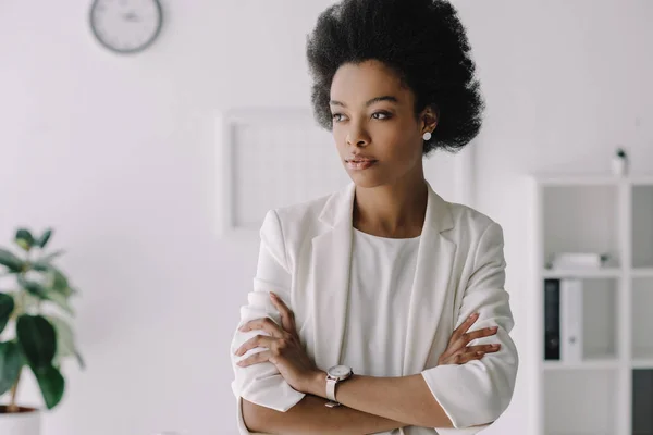 Atractiva mujer de negocios afroamericana de pie con los brazos cruzados en la oficina - foto de stock