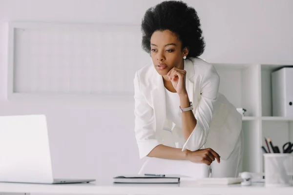 Atractiva mujer de negocios afroamericana apoyada en la mesa y mirando el ordenador portátil en la oficina - foto de stock