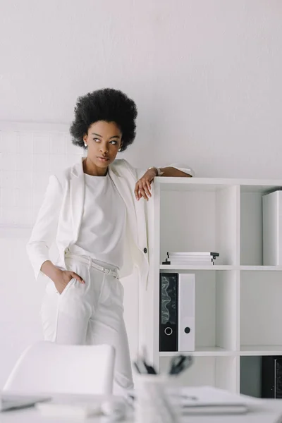 Hermosa mujer de negocios afroamericana apoyada en estantes y mirando hacia otro lado en la oficina - foto de stock