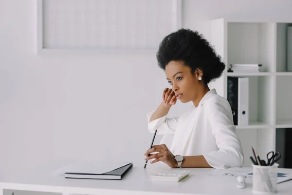Atractiva mujer de negocios afroamericana sentada en la mesa y mirando hacia otro lado en la oficina - foto de stock