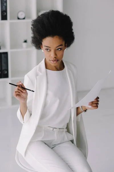 Atractiva mujer de negocios afroamericana sosteniendo documento y lápiz en la oficina - foto de stock