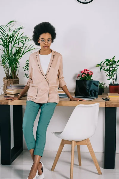 Atractiva mujer de negocios afroamericana apoyada en la mesa en la oficina - foto de stock