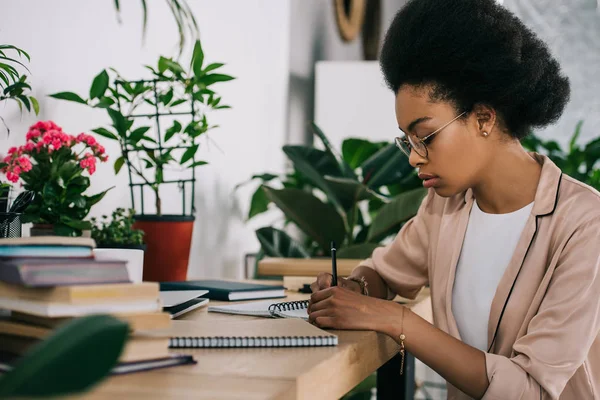 Visão lateral da atraente mulher de negócios afro-americana escrevendo algo para notebook no escritório — Fotografia de Stock