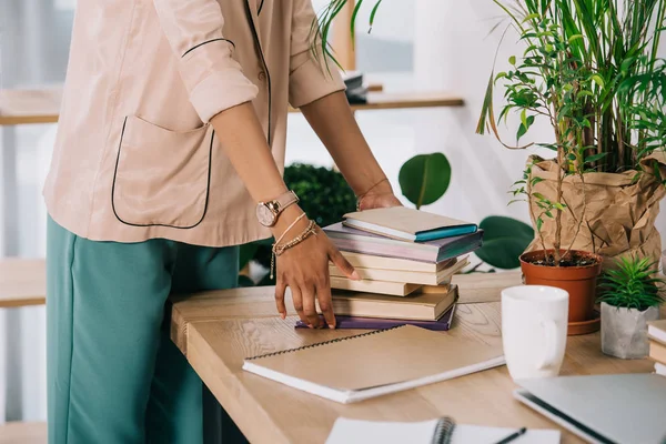 Imagen recortada de la mujer de negocios afroamericana poniendo libros en la mesa en la oficina - foto de stock