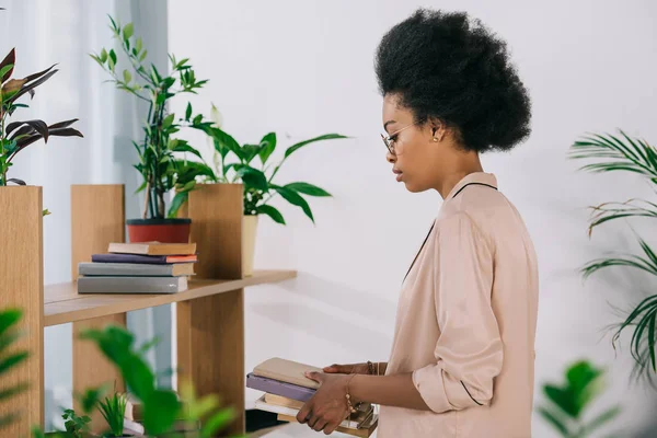 Visão lateral da atraente mulher de negócios afro-americana segurando livros no escritório — Fotografia de Stock