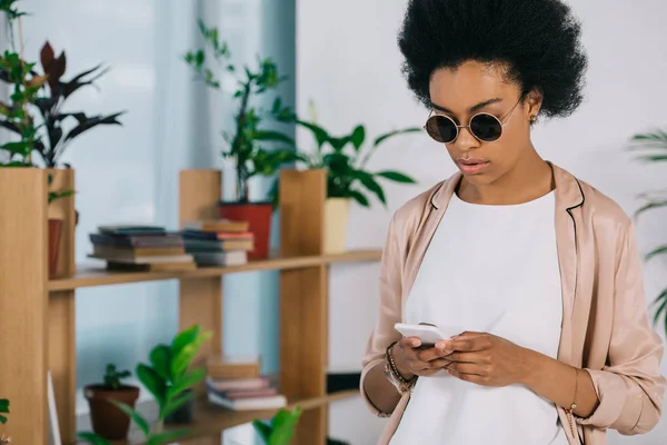 Attractive african american businesswoman in sunglasses using smartphone in office — Stock Photo