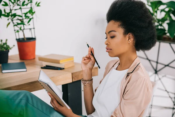 Séduisante femme d'affaires afro-américaine tenant un crayon et lisant des notes au bureau — Photo de stock