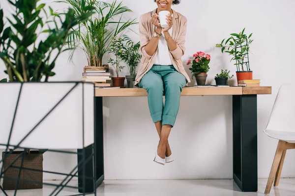 Imagen recortada de la mujer de negocios afroamericana sentada en la mesa con taza de café en la oficina - foto de stock