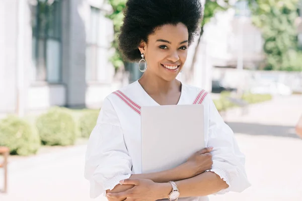 Atractiva mujer de negocios afroamericana sonriente de pie en la calle con el ordenador portátil - foto de stock