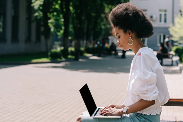 Side view of attractive african american businesswoman sitting on bench and using laptop on street — Stock Photo