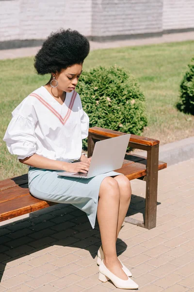 Attractive african american businesswoman sitting on bench and using laptop on street — Stock Photo