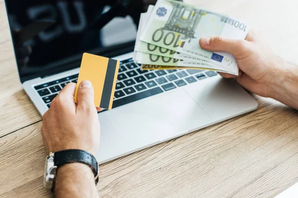 Cropped shot of person holding credit card and euro banknotes above laptop — Stock Photo